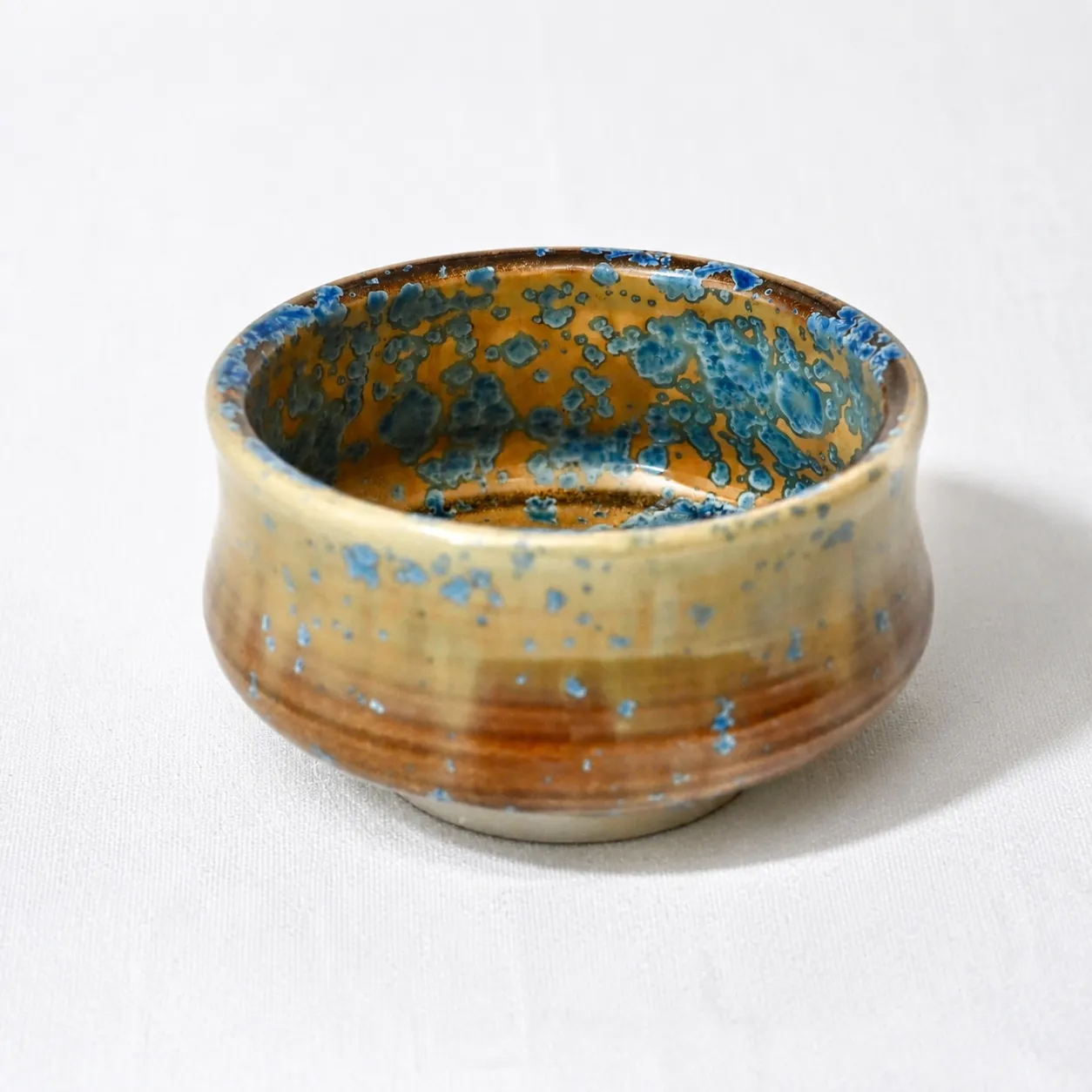 The bowl from the side, a low, wide form, amber glaze flecked with blue crystalline speckles over a warm foot
