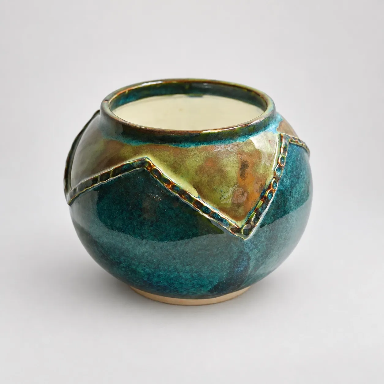 A squat round porcelain pot, deep teal-and-amber glaze with a raised zigzag band, and a cream interior