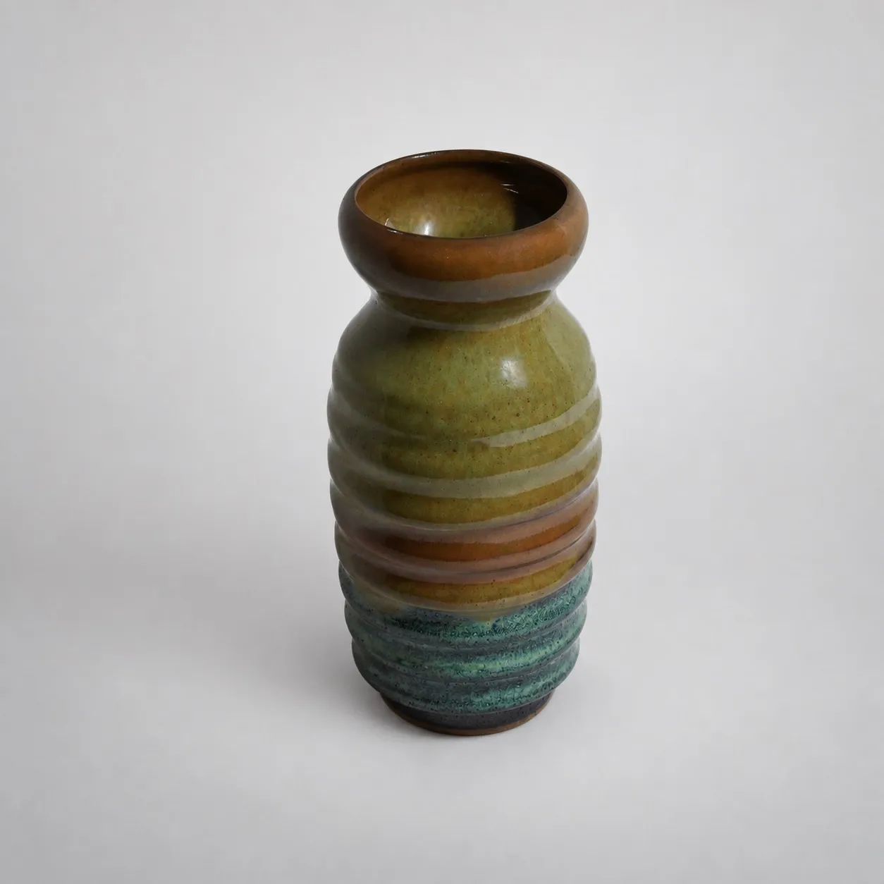 A tall ribbed stoneware vase with a rolled brown rim, olive-green above shifting to bands of teal-green below
