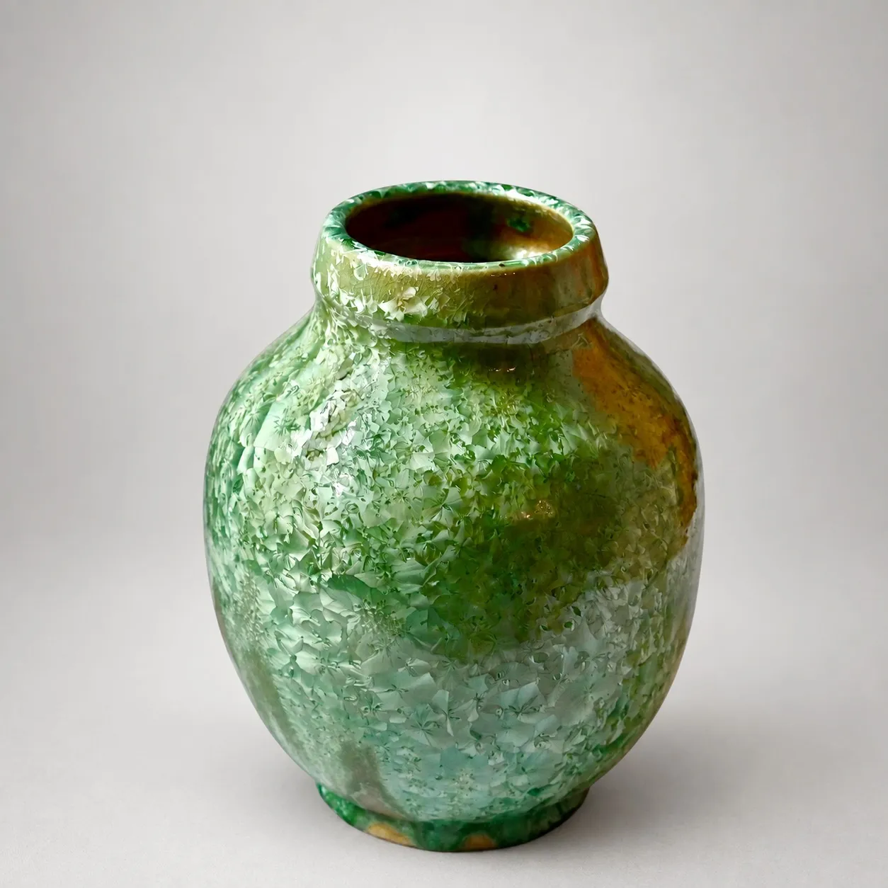 A rounded stoneware jar with a short rolled neck, green crystalline glaze mottling from olive to pale teal with amber breaking through at the foot