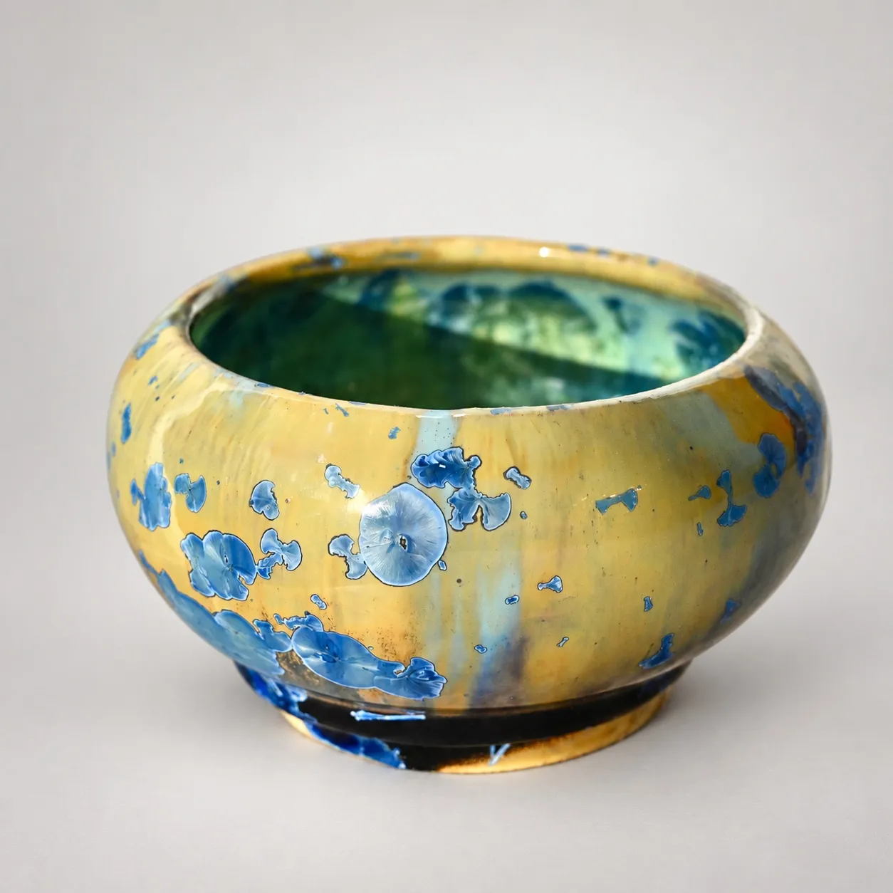 A small squat stoneware bowl, amber-beige glaze scattered with blue crystal blooms outside, a green interior showing at the rim