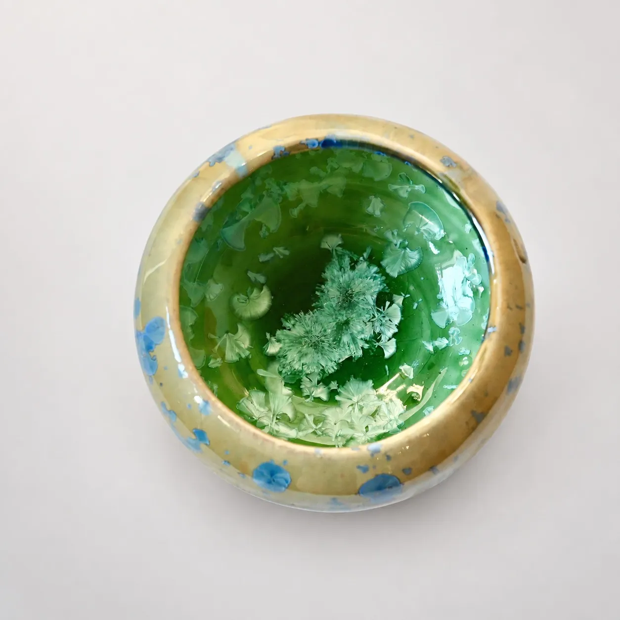 Looking down into the bowl, a deep green interior with pale green crystals blooming in the well, ringed by the amber-and-blue outer glaze