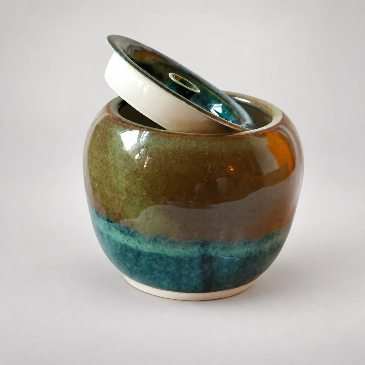 A round lidded porcelain jar, olive-and-amber glaze over a deep teal-blue band, its teal lid tilted ajar to show the pale interior