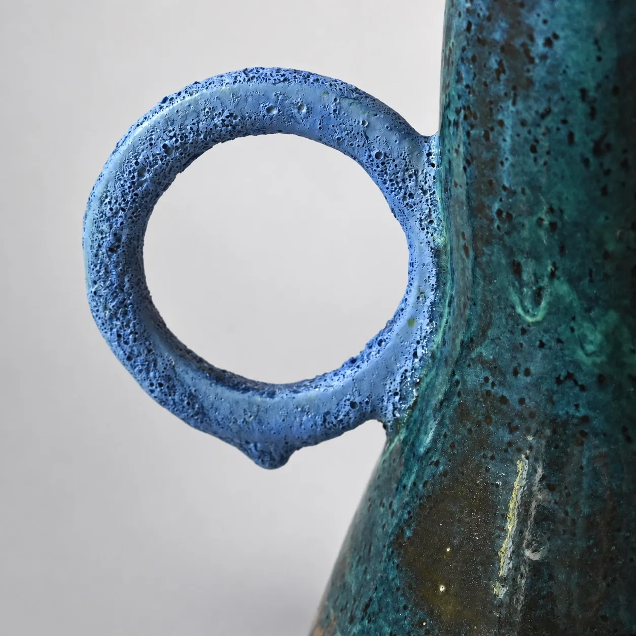 Close view of one ring handle, its pumice-textured volcanic blue glaze against the speckled teal body