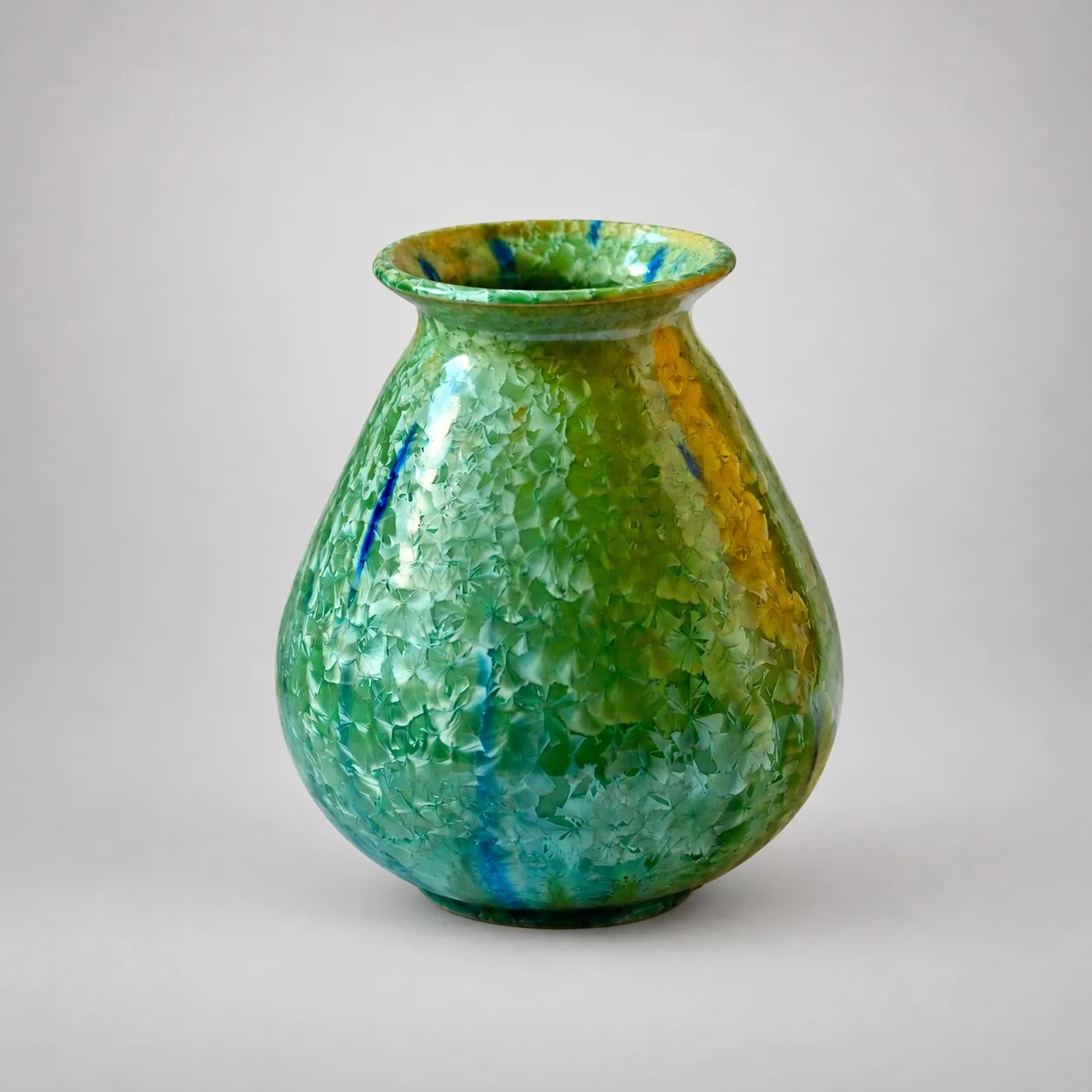 A teardrop-shaped stoneware vase with a flared rim, dense green crystalline blooms over the whole body with cobalt-blue streaks running down and amber at one side