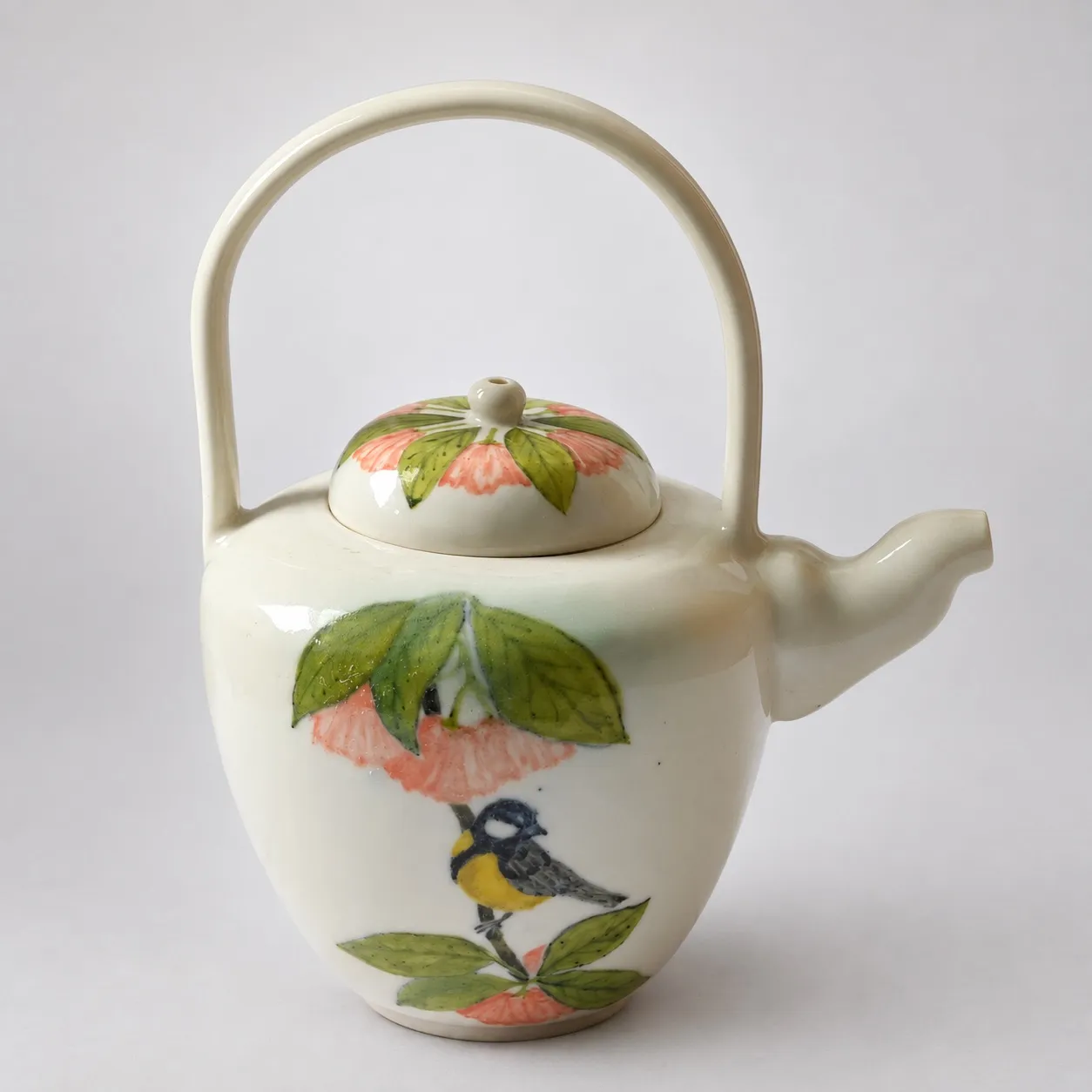 A cream porcelain teapot with an overhead loop handle and a short spout, hand-painted with a great tit perched among green leaves and coral blossoms
