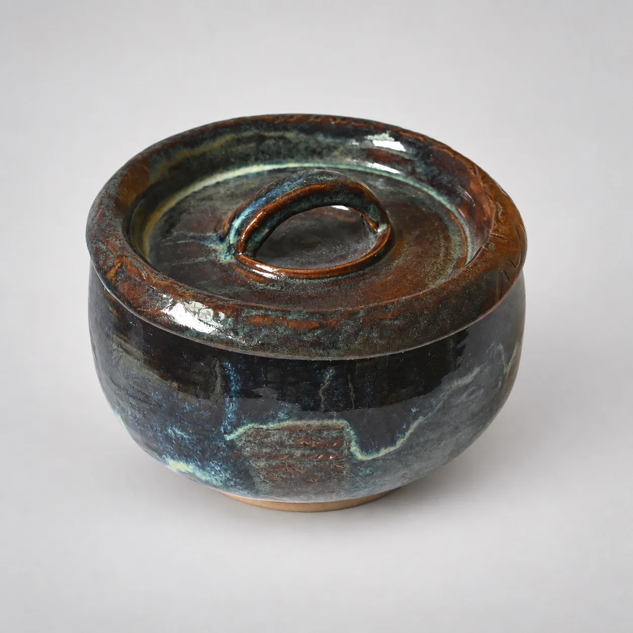 A squat lidded stoneware jar, dark blue-black rutile glaze breaking to amber, with a ribbon-loop knob and an incised band around the lid rim