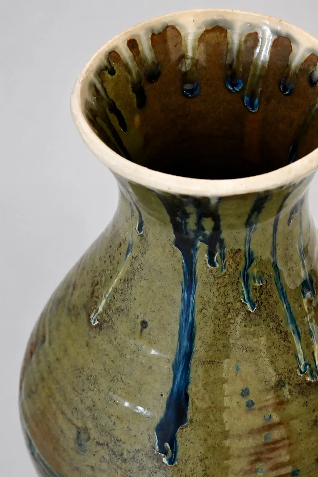 The neck and shoulder, blue drips hanging from the rim into a warm amber interior, blue runs trailing down the olive ground