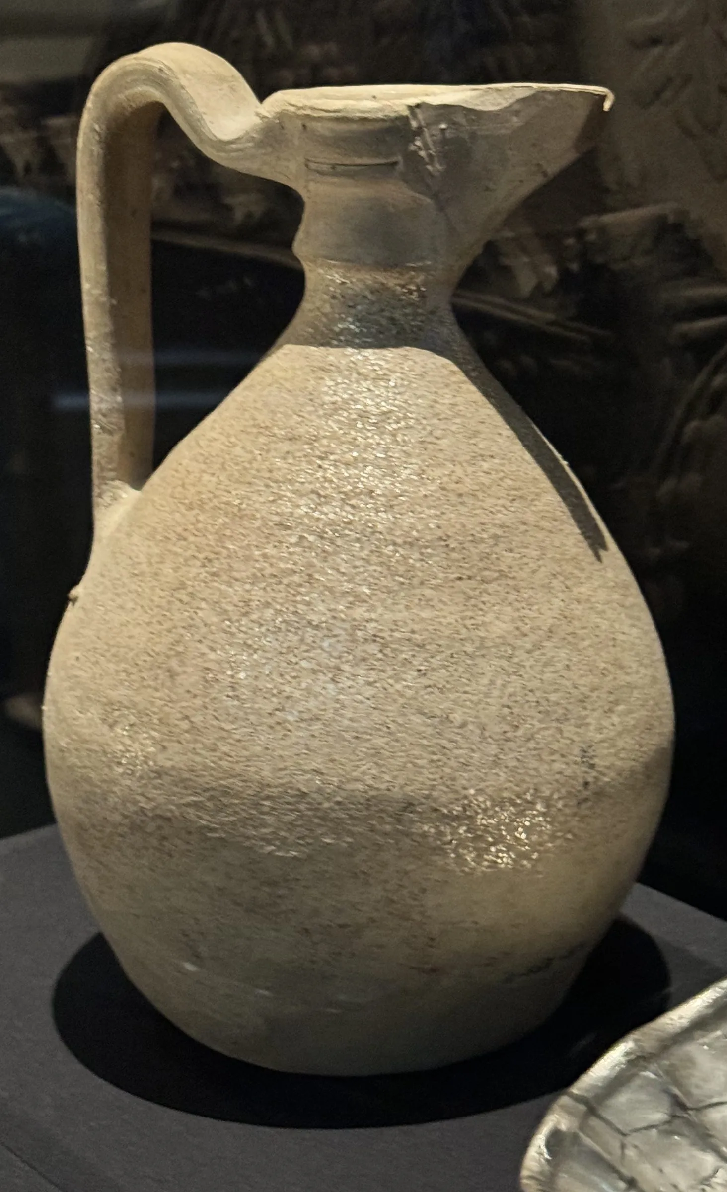 A pale grey clay jug in a museum case: a full pear-shaped body drawn in to a short neck, a pinched pouring lip at the front and a tall strap handle behind, the bare surface flecked and glittering with mica.
