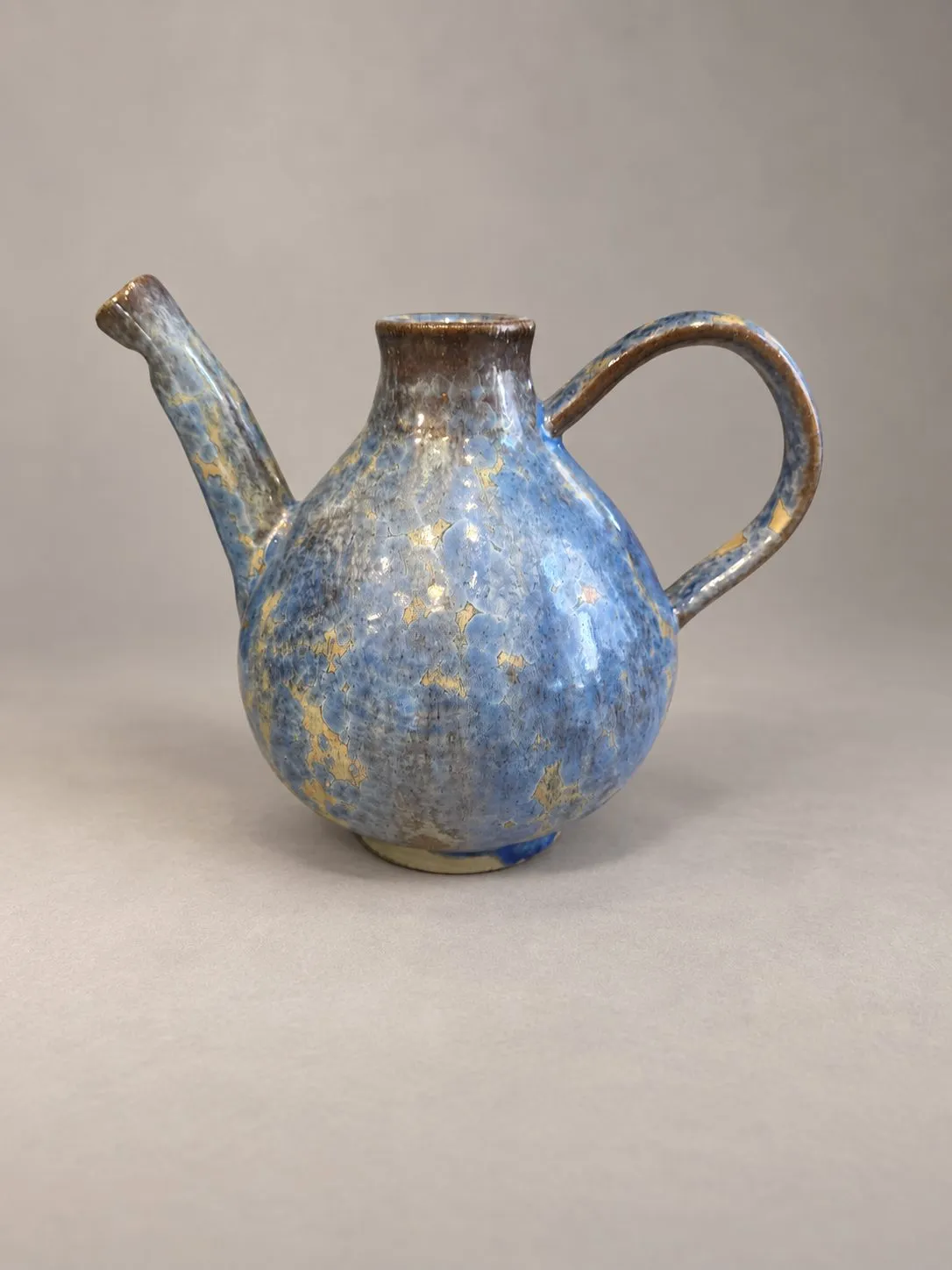 A stoneware coffee pot by Igor Krol: a full pear-shaped body on a small foot, a short collared neck, a long straight spout rising from the lower body and a high looping handle, in mottled blue breaking to ochre and brown.