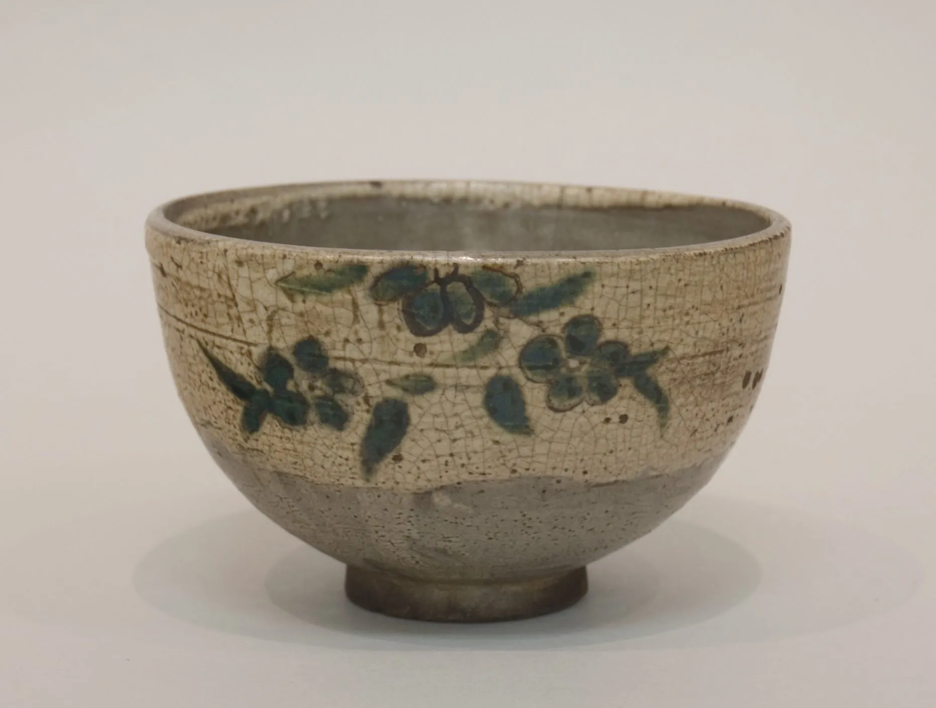 A Japanese tea bowl: a rounded hemisphere sitting low on a small foot, wider than it is tall, the upper wall carrying a sparse painted branch of leaves and blossom, the lower wall left bare.