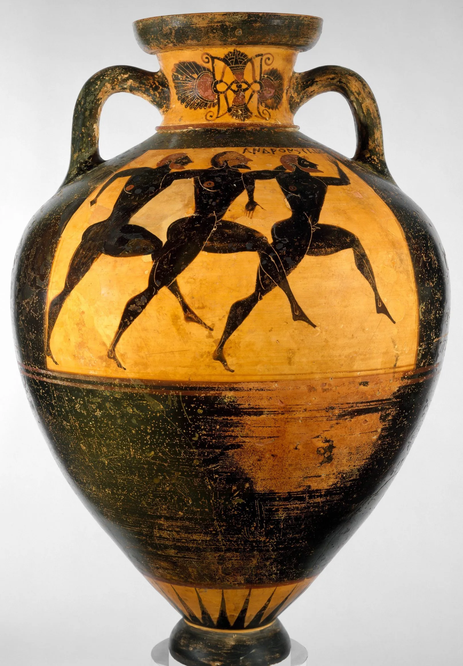 A tall Greek amphora: a broad shoulder narrowing to a small foot, a short neck with a heavy rolled lip, two handles, and three runners painted in black striding across the belly.