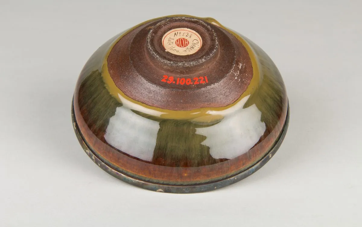 The bowl turned over: a small turned foot ring of bare brown clay cut in shallow steps, where the glaze stops in a thick roll, carrying an old collector’s paper label and a painted accession number.