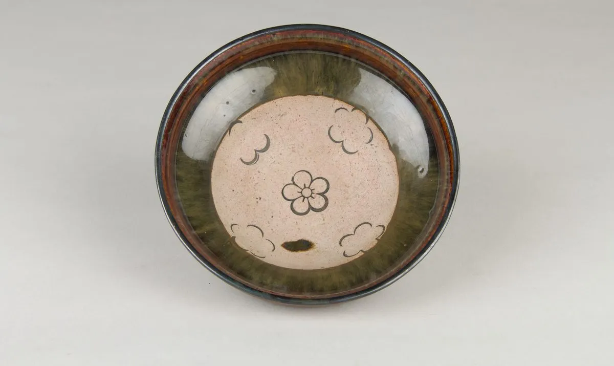 Looking down into the bowl: a broad unglazed disc in the centre of the floor, drawn with a five-petalled plum flower and small clouds, ringed by the darker glazed wall.