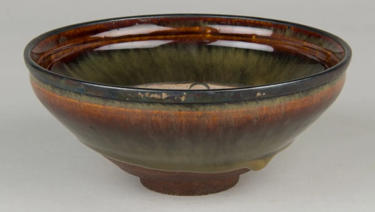 A shallow Chinese bowl seen from the side: a curved wall rising from a small turned foot to a rim bound in a thin metal band, the surface running from dark olive to russet brown.