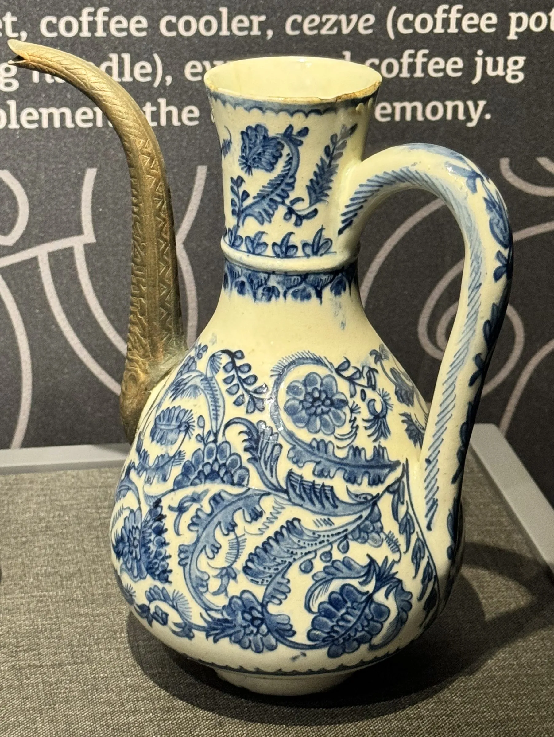 An Ottoman coffee jug in a museum case: a pear-shaped body under a long neck with a flaring mouth, painted all over in blue with leaves and flowers, a high looping handle on one side and a long curved brass spout on the other.