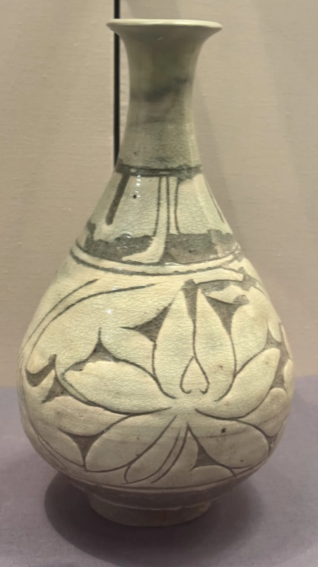 A pale grey-green Chinese bottle in a museum case: a rounded pear body carrying a large carved lotus, drawn in to a long neck that opens at the top into a small dish-shaped mouth.