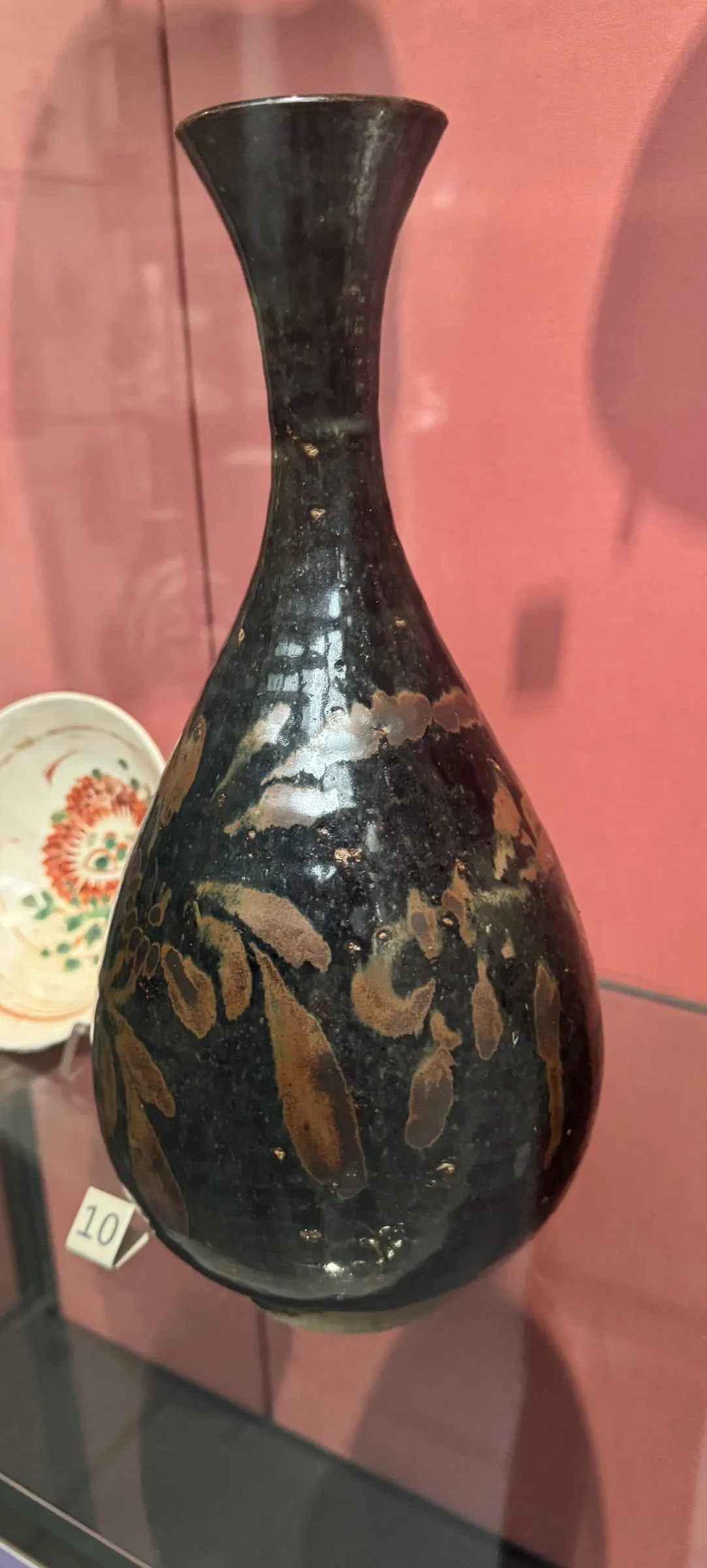 A Chinese bottle in a museum case: a full pear-shaped body low down, a long neck rising from it, and a mouth flaring out at the top. The dark surface is scattered with russet leaf-shaped markings.